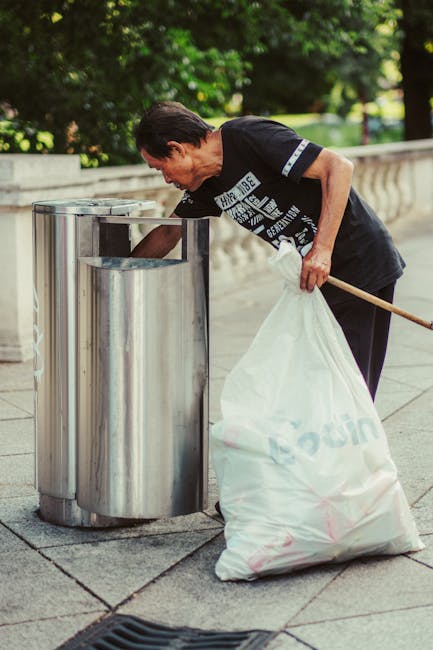 A middle-aged man with dark hair, wearing a black T-shirt with white text and dark pants, is seen discarding waste into a modern stainless steel recycling bin located on a paved pedestrian walkway. He is bending slightly forward and holding a large white bag filled with rubbish in his left hand, which rests against his hip, while his right hand is actively placing waste into the top opening of the bin. The bin has a sleek, cylindrical design with multiple compartments, set against a backdrop of lush green trees and a stone balustrade, suggesting an outdoor, urban park or public space environment. The natural daylight illuminates the scene, highlighting the textured metallic surface of the bin and the fabric of the man's clothing. This image exemplifies independent waste disposal, aligning with private rubbish collection services such as those offered by Rubbish Collection Sutton, emphasizing on-site rubbish handling in public or communal areas, with a focus on responsible waste management and recycling efforts.