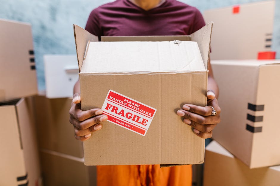 A person wearing a maroon top and orange skirt holds an open cardboard box with both hands, prominently displaying a red and white 'Fragile' handle with the message 'Please handle with care' printed on it. The individual is surrounded by other sealed cardboard boxes stacked and arranged on a flat surface, likely in a warehouse or storage area. The background features a slightly blurred wall with a textured, light blue surface. The boxes vary in size and are made of brown, corrugated cardboard with black tape securing some edges. The lighting is even and neutral, highlighting the textures of the boxes' surfaces and the details of the 'Fragile' sticker. This scene reflects themes of careful handling and packaging, relevant to private waste disposal services by companies like Rubbish Collection Sutton involved in managing waste removal, packaging, or on-site clearance services in a professional context.