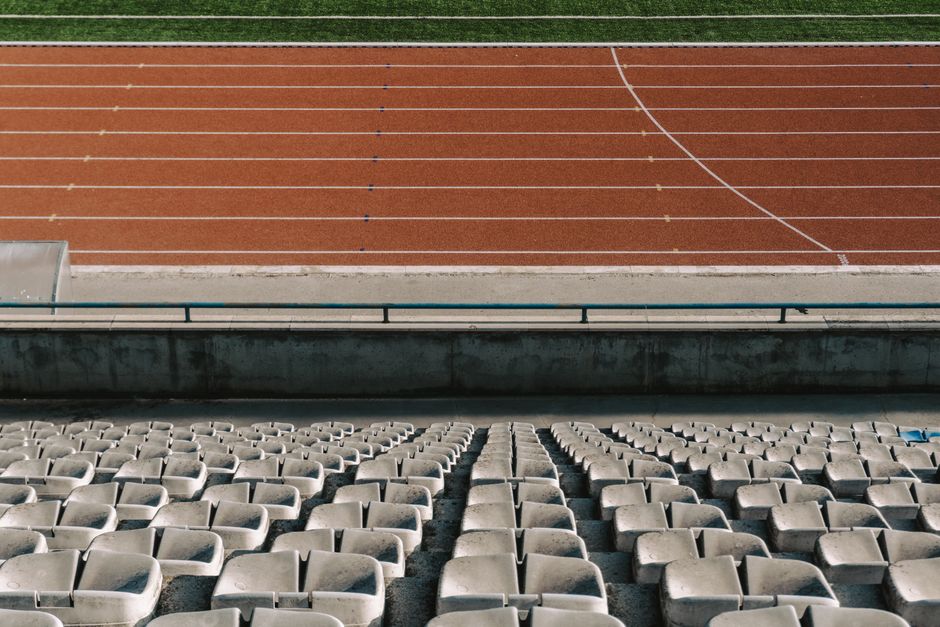 The image shows the view from a stadium bleacher seating area overlooking a running track. In the foreground, there are multiple rows of weathered, light gray plastic seats arranged in a curved formation, with some showing signs of dirt and wear. The seats are closely spaced with a slight gap between each, and are mounted on metal frames. Behind the seats, a concrete barrier runs horizontally across the image, separating the seating area from the track. The running track itself is reddish-brown with white lane markings, and appears clean and well-maintained, with distinct lines and curves indicating multiple lanes. The background includes a narrow strip of green grass along the edge of the track, adding a natural contrast to the synthetic surface. The overall lighting suggests a clear day with natural sunlight illuminating the scene evenly, highlighting the textures of the plastic seats and the precise markings on the track. This setting reflects a typical sports stadium environment, where seating and track are designed for athletic events and training activities.