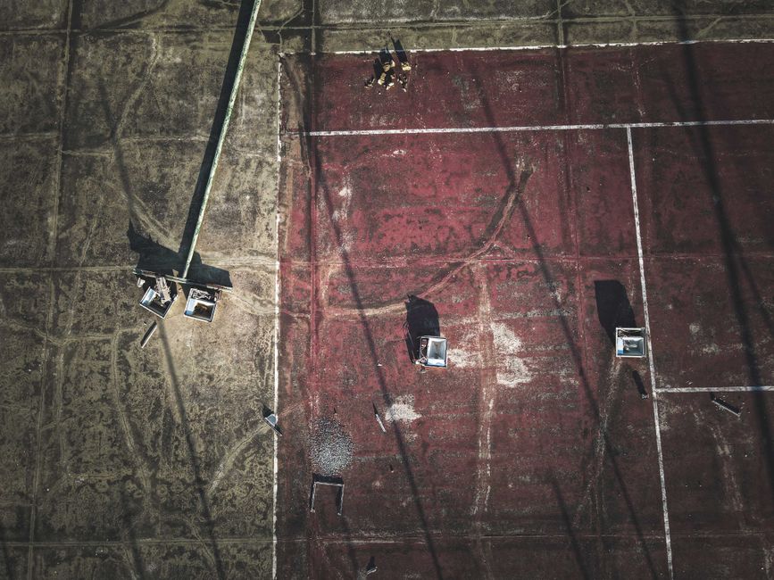 An aerial view of an outdoor sports court surface, divided into two sections: the left side features a worn, grayish concrete area with visible tire marks and scuff marks, while the right side displays a faded red surface with dirt and stains. Several temporary floodlights with metal frames and rectangular light fixtures are positioned on the court, casting long shadows across the surface. A tall, metal pole supporting a large floodlight is situated on the left side, with its shadow stretching across the concrete area. The scene appears to be part of an independent or private sports facility, with no surrounding infrastructure visible. The overall atmosphere is subdued, with natural and artificial lighting highlighting the textured surfaces and the remnants of previous activity, suggesting recent or ongoing maintenance or cleaning operations related to waste or debris removal, possibly carried out by Rubbish Collection Sutton in the context of site clearance or waste management work.