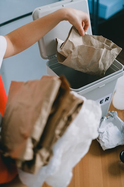 A person discards crumpled brown paper bags into a white rubbish bin with a black lid, positioned on a wooden surface. The person's hand, visible in the image, holds one of the paper bags, which is partially open and appears to contain waste. The bin is situated next to various other crumpled paper bags and waste items, suggesting a household or small-scale waste disposal setting. The background includes a grey wall and a window, providing natural lighting that illuminates the scene clearly. The image emphasizes the process of private or alternative rubbish disposal, often handled by waste management services such as Rubbish Collection Sutton, which offers on-site clearance and independent waste collection options for households looking to manage their rubbish separately from local authority services.