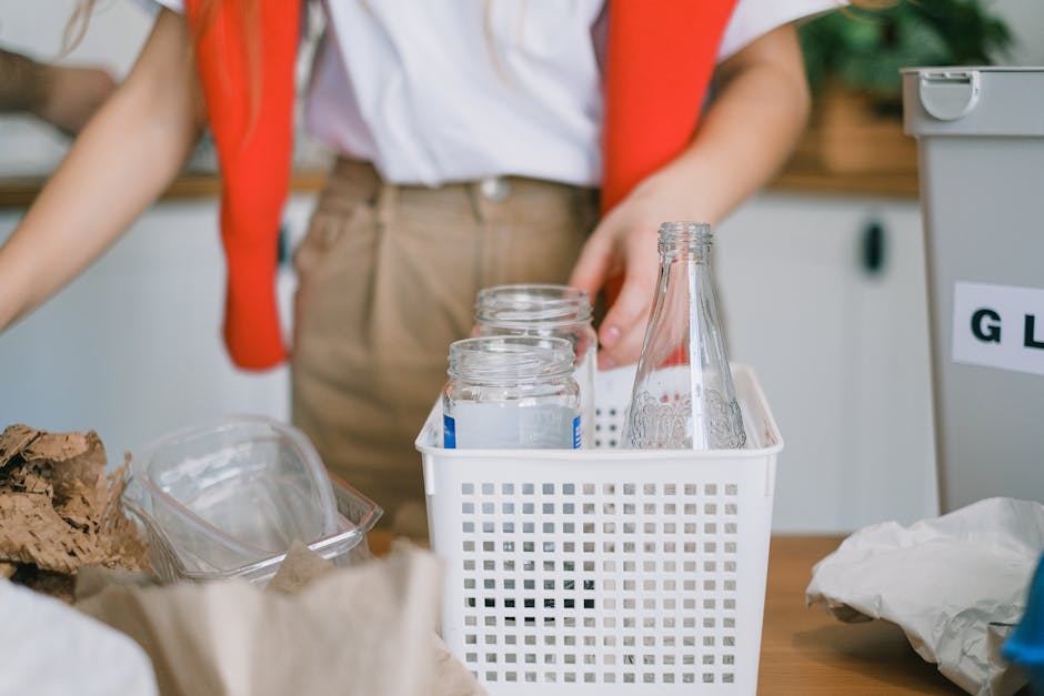 The image shows a close-up of a person’s midsection, wearing a white shirt with a red scarf draped over their shoulders and beige trousers, standing behind a cluttered workspace or kitchen counter. In the foreground, there is a white plastic bin with a grid pattern, containing several empty glass jars with metal screw caps and a clear glass bottle, all clean and neatly arranged. To the left, there are crumpled brown paper and some transparent plastic containers, while on the right side, part of a gray rubbish collection bin with a white label visible, indicating it is used for general waste. The environment suggests an indoor setting, possibly a recycling or waste sorting area, where waste items like glass bottles and paper are temporarily stored before disposal. The overall scene emphasizes an organized approach to waste segregation, aligning with private waste handling or rubbish removal services like those provided by Rubbish Collection Sutton, often utilized for household or commercial rubbish clearance.