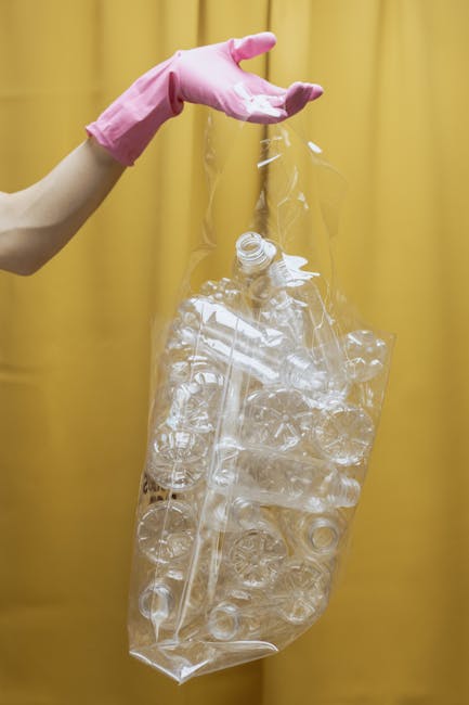 A person discards crumpled brown paper bags into a white rubbish bin with a black lid, positioned on a wooden surface. The person's hand, visible in the image, holds one of the paper bags, which is partially open and appears to contain waste. The bin is situated next to various other crumpled paper bags and waste items, suggesting a household or small-scale waste disposal setting. The background includes a grey wall and a window, providing natural lighting that illuminates the scene clearly. The image emphasizes the process of private or alternative rubbish disposal, often handled by waste management services such as Rubbish Collection Sutton, which offers on-site clearance and independent waste collection options for households looking to manage their rubbish separately from local authority services.